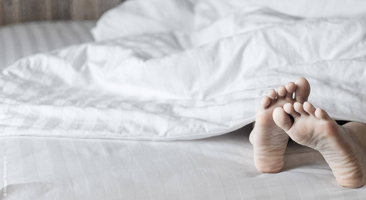 Foto: Auf einem weißen Spannbettlaken liegt eine weiße Bettdecke. Ein Paar Füße schaut darunter hervor.