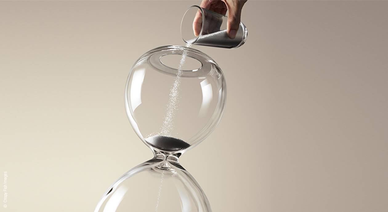 Foto: Eine Sanduhr, die oben offen ist. Eine Hand schüttet mit einem Glas mehr Sand ins Stundenglas.