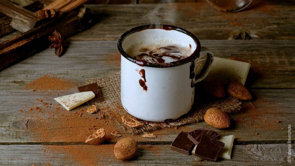 Foto: Auf einem Holztisch steht eine dampfende Tasse Kakao. Daneben liegen Mandeln und Schokosplitter
