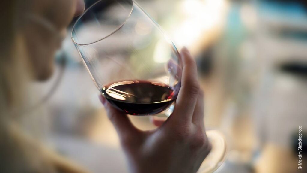 Foto: Eine Person wird von hinten gezeigt, wie sie an einem Glas Rotwein nippt.