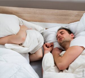 Foto: Ein Paar liegt im Doppelbett. Eine Person drückt sich ein Kissen auf den Kopf. Die andere schläft und scheint dabei zu schnarchen.