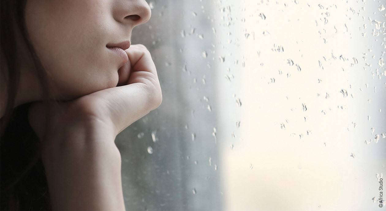 Foto: Eine Person schaut, das Kinn auf die Hand gestützt, gedankenversunken aus dem Fenster, an der Scheibe Regentropfen.