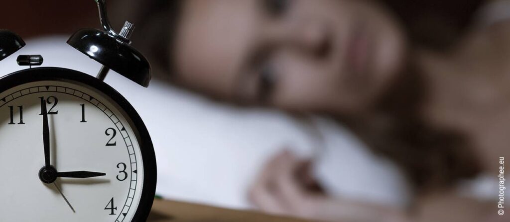 Symbolbild Schlafmangel durch Winterdepression: Ein altmodischer Wecker mit Zeigern, dahinter liegt eine Person wach im Bett.