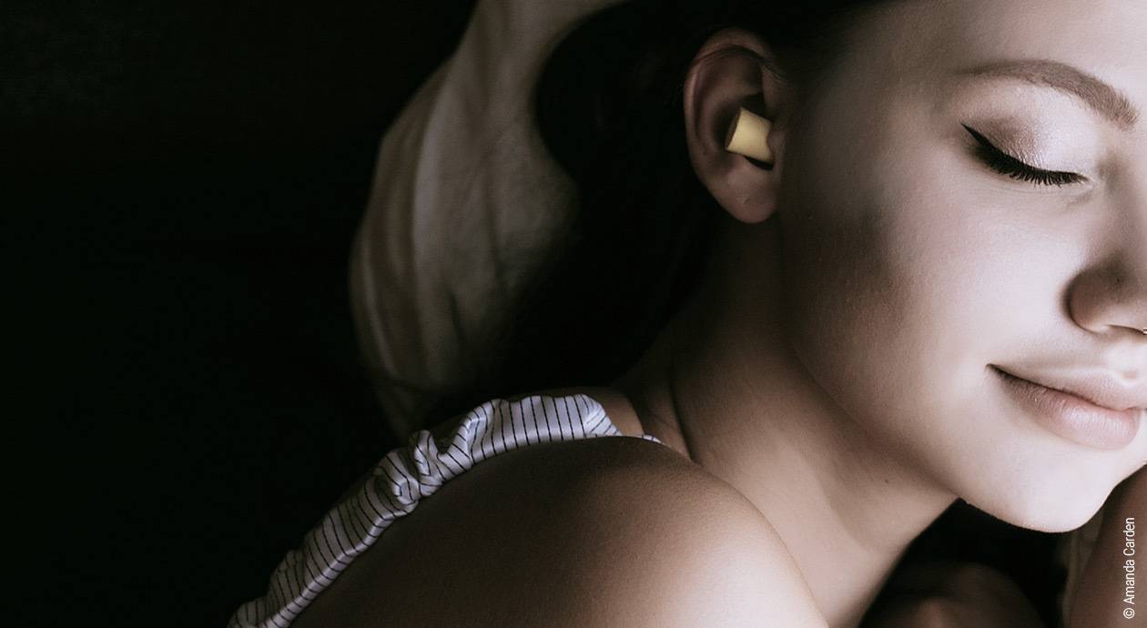 Foto: Eine Frau liegt mit geschlossenen Augen in Dunkelheit. In ihrem Ohr steckt ein Ohrstöpsel.