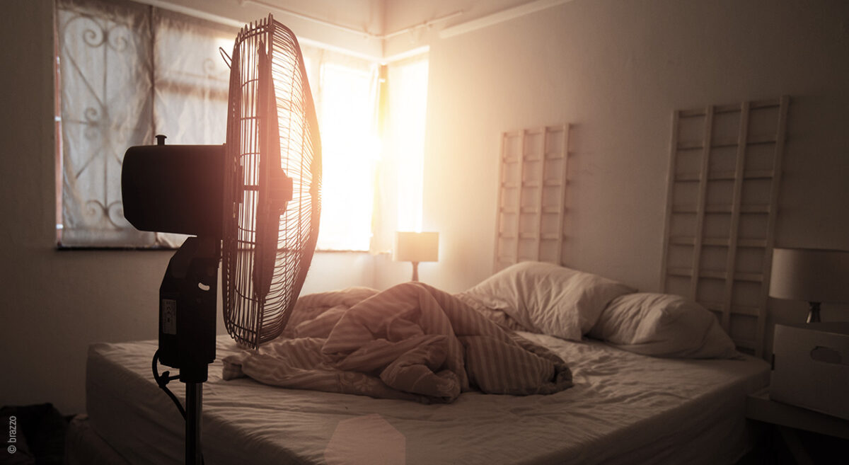 Foto: Die Sonne scheint durch die hellen Vorhänge eines abgedunkelten Schlafzimmers; im Vordergrund ein Ventilator.