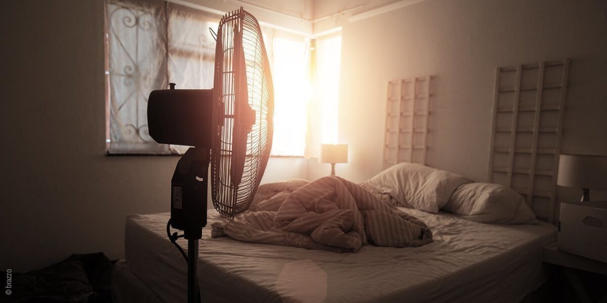 Foto: Die Sonne scheint durch die hellen Vorhänge eines abgedunkelten Schlafzimmers; im Vordergrund ein Ventilator.