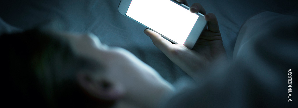 Das Gesicht einer Person schaut nachts liegend im Bett auf das grelle Display des Smartphones, welches sie in der Hand hält.