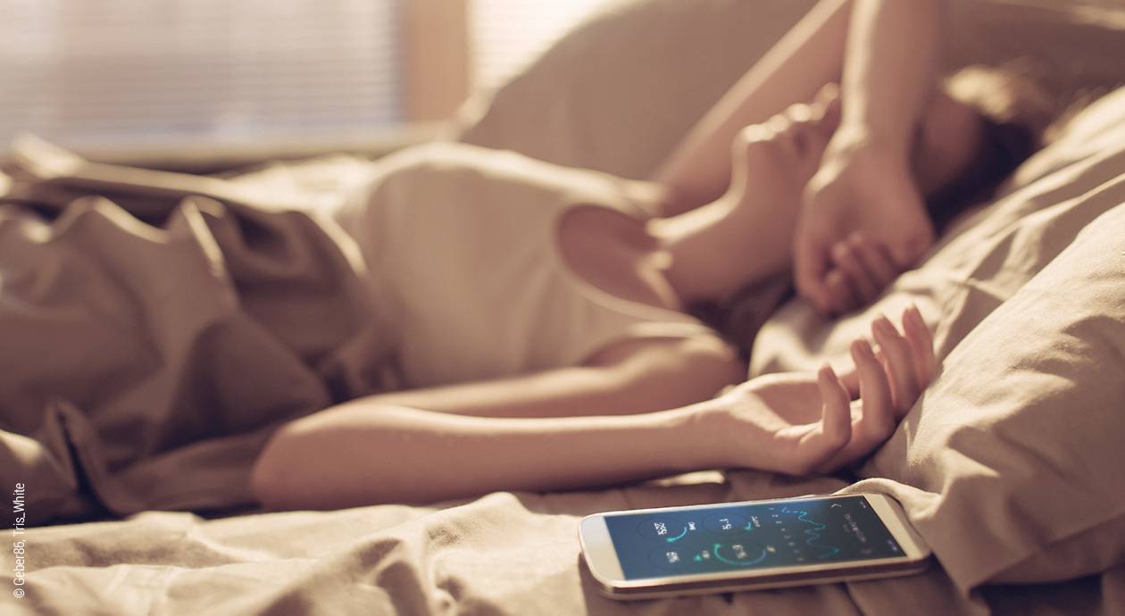Foto: Eine Frau wacht morgens in ihrem Bett auf. Daneben liegt ihr Smartphone im Bett.