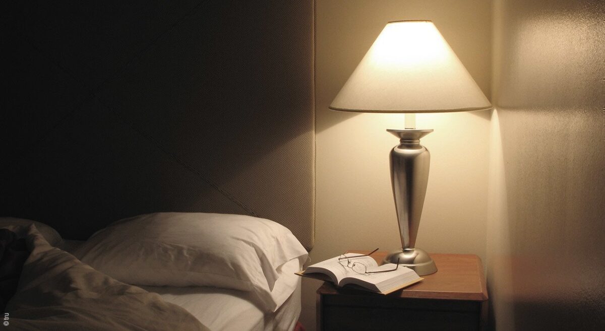 Foto: Auf einem Nachttisch neben einem Bett liegt ein Buch und eine angeschaltete Nachttischlampe.