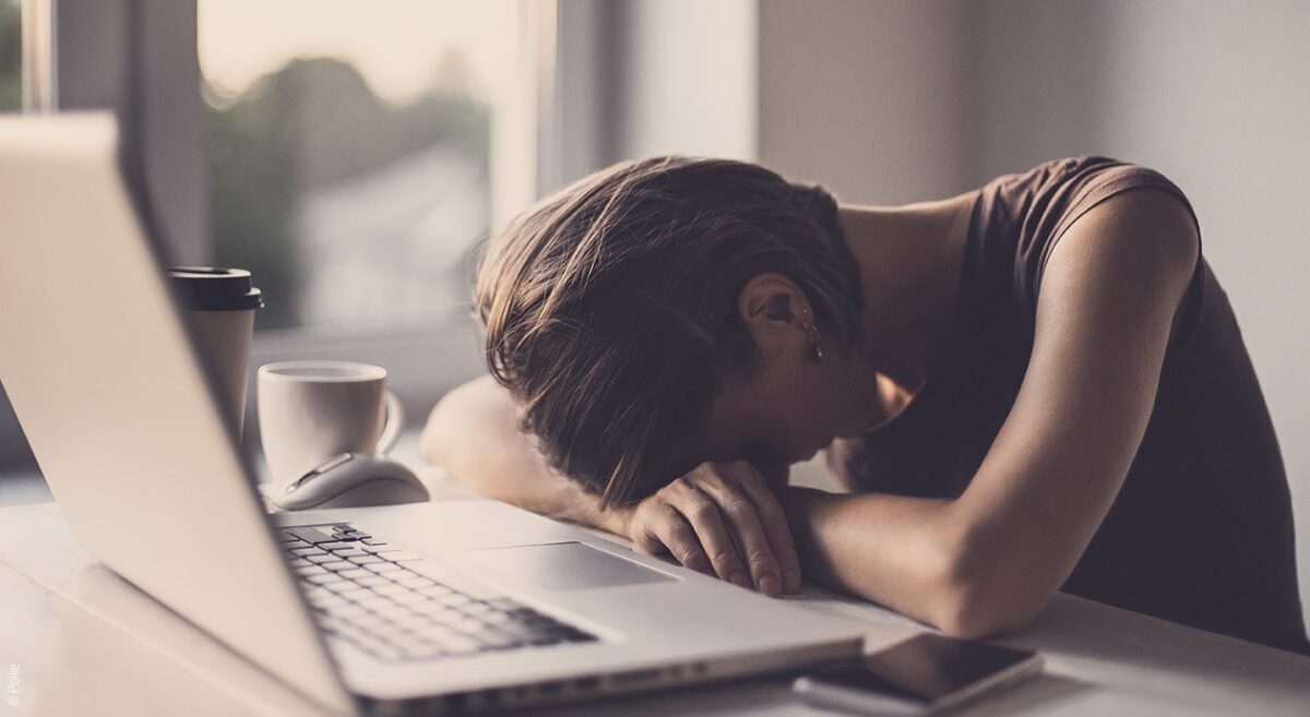 Symbolfoto Narkolepsie: Eine vor einem Laptop sitzende Person legt erschöpft ihren Kopf in ihre verschränkten Arme.
