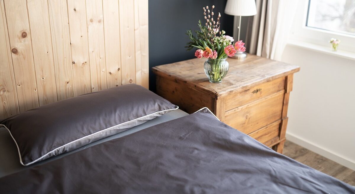 Foto: Die BODYGUARD Satin Bettwäsche auf einem Bett, daneben ein steht ein Beistelltisch aus Holz. Auf dem Beistelltisch stehen Blumen.