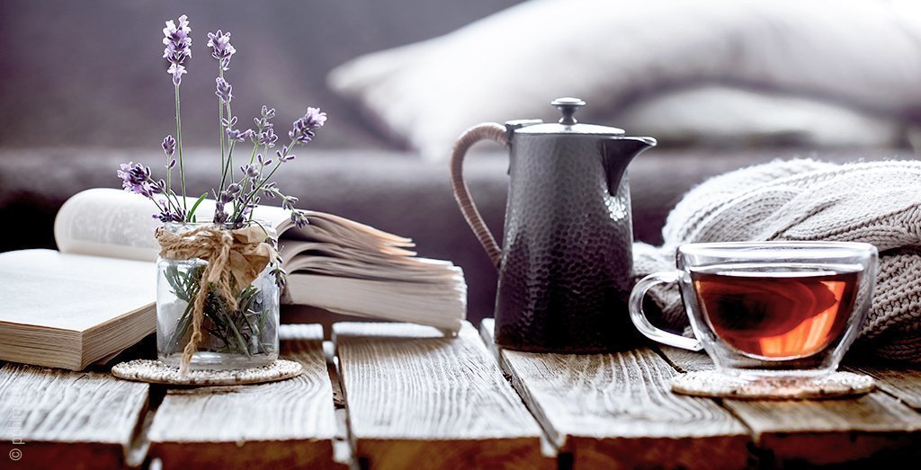 Foto: Ein Holztisch; darauf befindet sich ein Buch, ein kleiner Strauß Blumen, eine Teekanne und ein gefüllte Tasse Tee – eine Ruhe-Routine gehört zur Schlafhygiene.