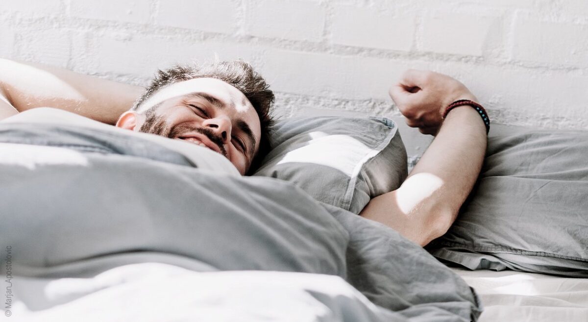 Foto: Symbolbild zur Schlafhygiene: Ein Mann liegt in einem Bett; er streckt sich und sieht sichtlich erholt aus.