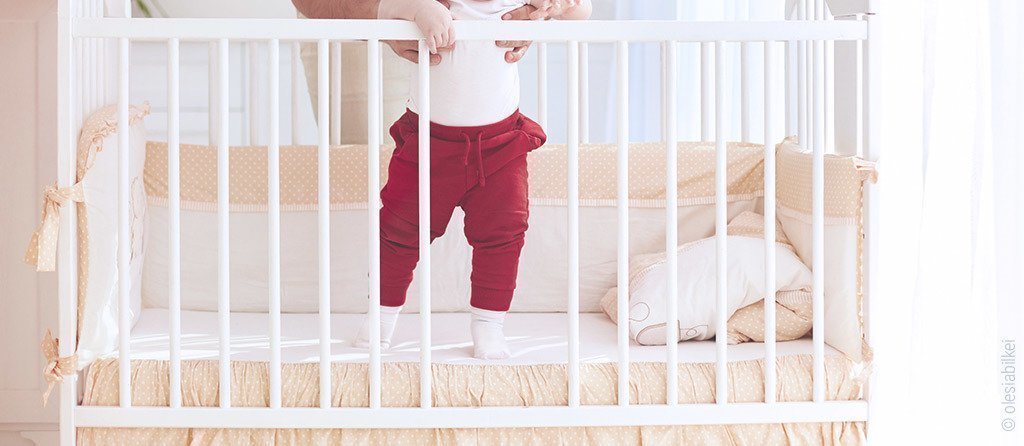 Foto: Ein Kind steht im Gitterbett aus dem zwei Schlupfsprossen entfernt wurden.