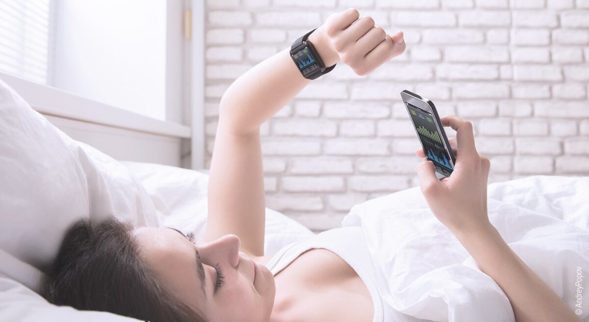 Foto: Eine Frau liegt im Bett und schaut auf ihre Smartwatch am linken Arm und in ihr Smartphone in der rechten Hand.