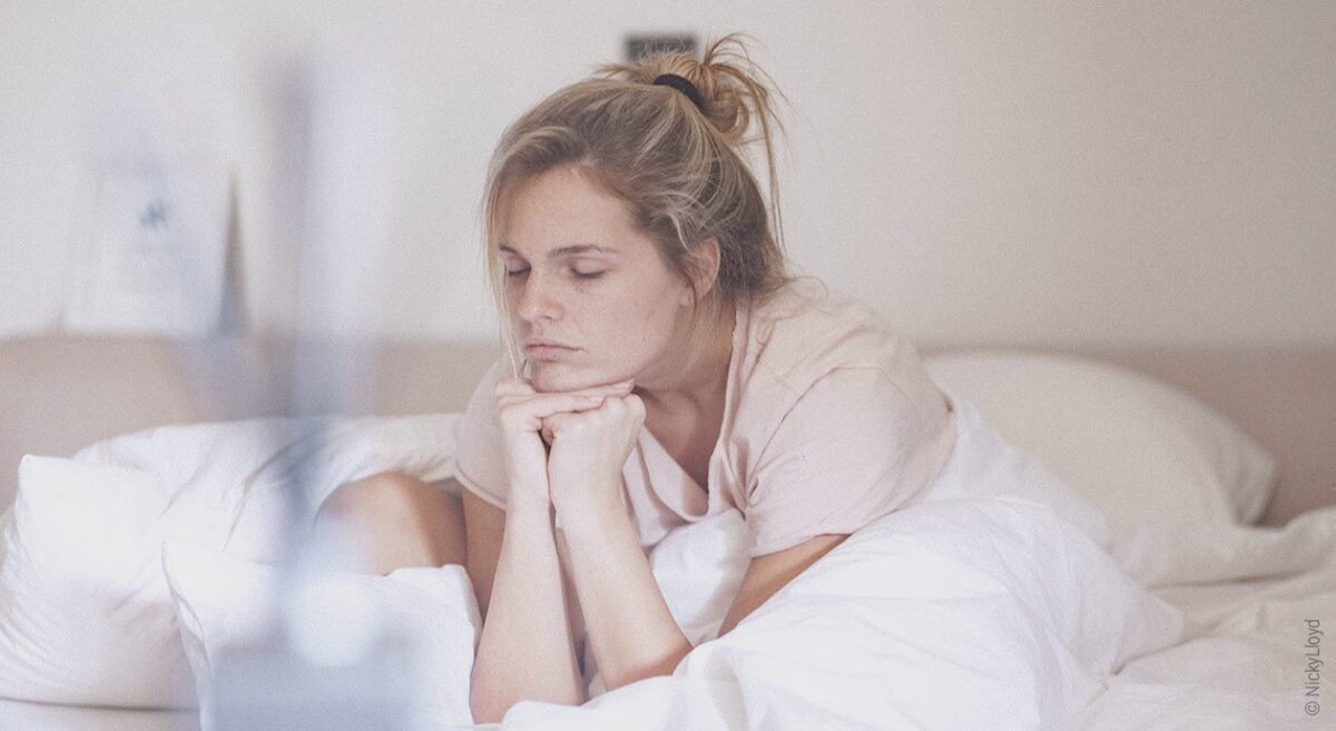 Symbolfoto Chronische Müdigkeit: Eine Frau sitzt im Bett, hat die Augen zu und stützt den Kopf auf den Händen ab.