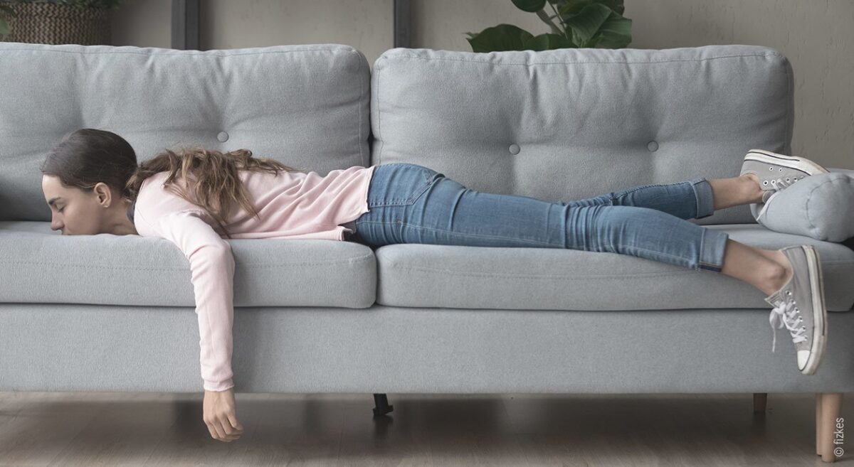 Foto: Eine junge Frau liegt lang ausgestreckt bäuchlings auf der Couch. Der linke Arm hängt vom Sofa runter.