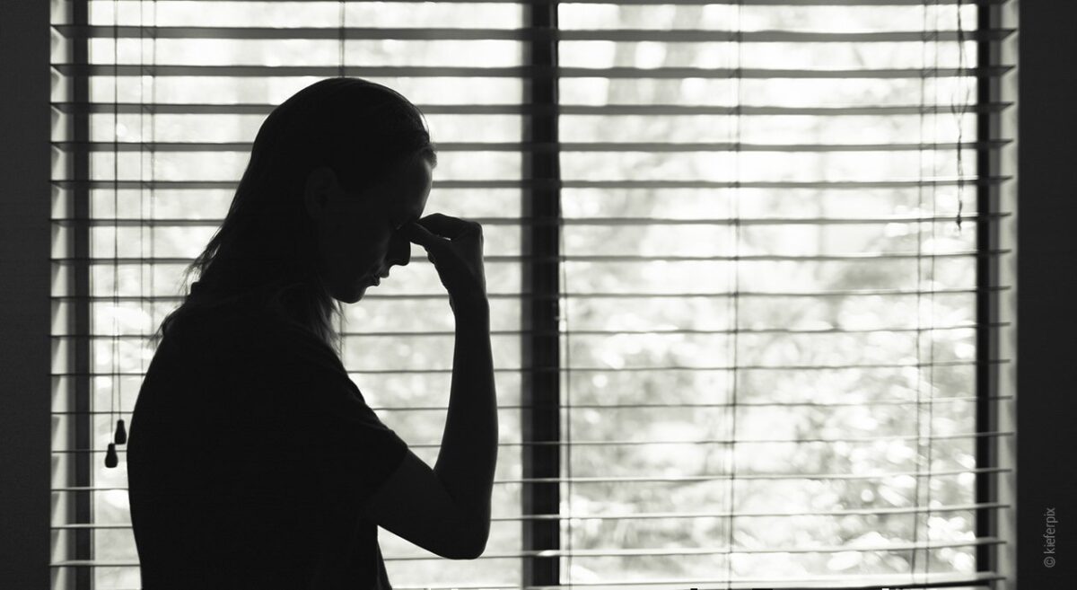 Symbolfoto Fatigue Syndrom: Die dunkle Silhouette einer Person steht vor einer aufgeklappten Jalousie. Sie fasst sich an den leicht nach vorne gebeugten Kopf.