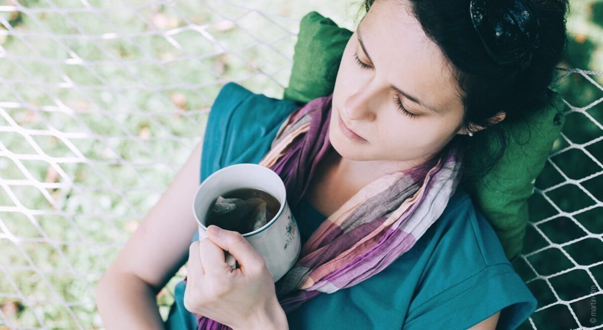 Foto: Eine Person liegt mit geschlossenen Augen in einer Hängematte. In einer Hand hält sie eine Tasse Tee.