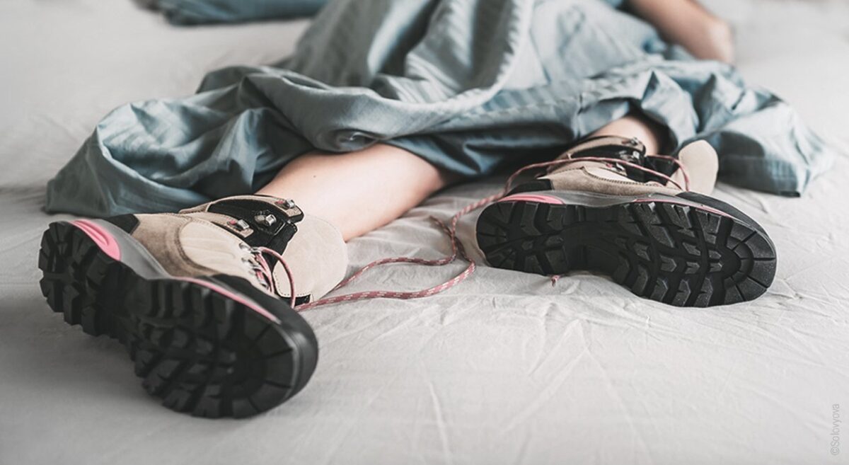 Symbolfoto für Restless-Legs-Syndrom: Fokus auf Wanderschuhe, die eine im Bett liegende Person trägt.