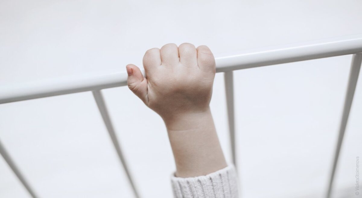 Foto: Eine Babyhand greift nach oben an die Stange eines Bettgitter aus Metall