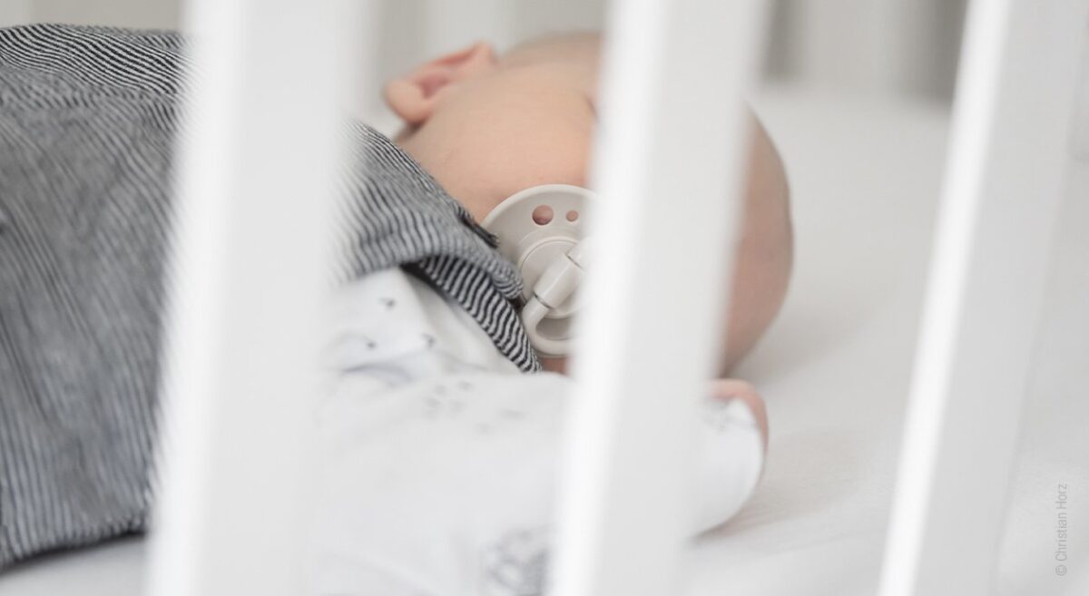 Symbolfoto Babyschlafsack: Hinter den Holzleisten eines Gitterbettes liegt ein Baby mit Schnuller in Rückenlage.