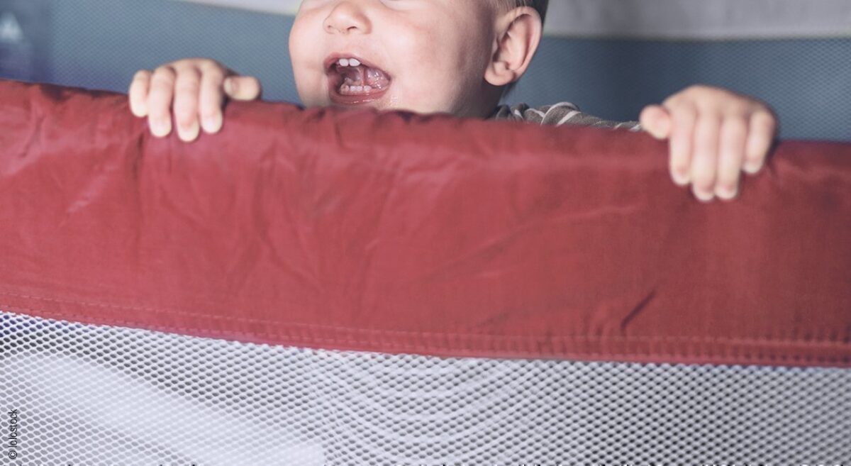 Foto: Ein Kleinkind steht aufrecht in einem Babyreisebett und hält sich lachend am Netzgitter fest.