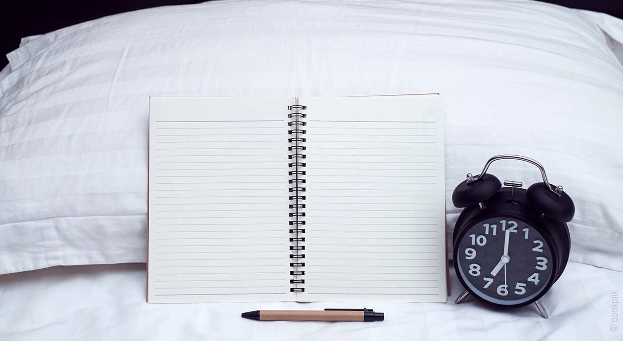 Foto: Ein offenes Notizbuch angelehnt an einem weißen Kissen, davor liegt ein Stift. Neben dem Notizbuch steht ein schwarzer Wecker