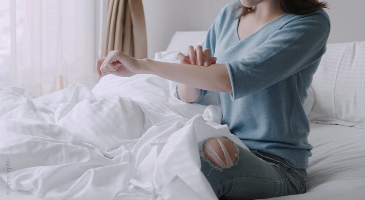 Foto: Eine Person sitzt im Bett und tastet ihren Unterarm ab.