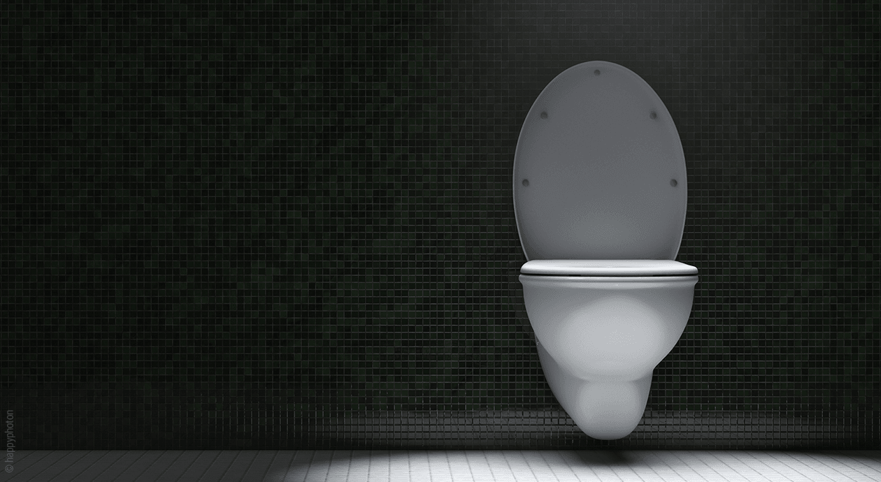Titelbild: Eine spärlich beleuchtete weiße Toilette ist in einem ansonsten dunklen Raum an einer schwarzgefliesten Wand angebracht.
