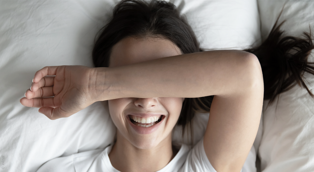 Foto: Eine Person liegt im Bett, hält sich den Unterarm über die Augen und lacht.