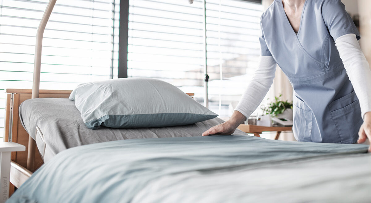 Foto: Eine Person in medizinischer Arbeitskleidung richtet das Bettzeug auf einem Pflegebett her.