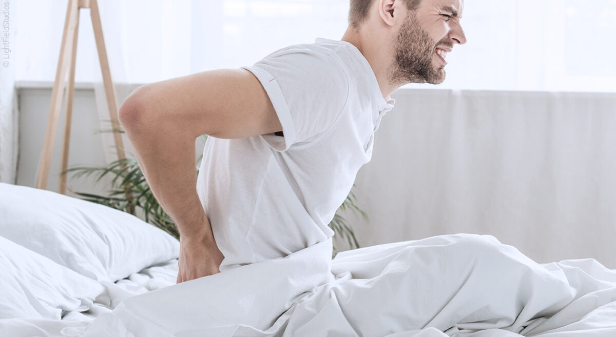 Titelbild zu Rückenschmerzen nach dem Schlafen: Eine Person sitzt im Bett und hält sich mit schmerzverzerrtem Gesicht den unteren Rücken.