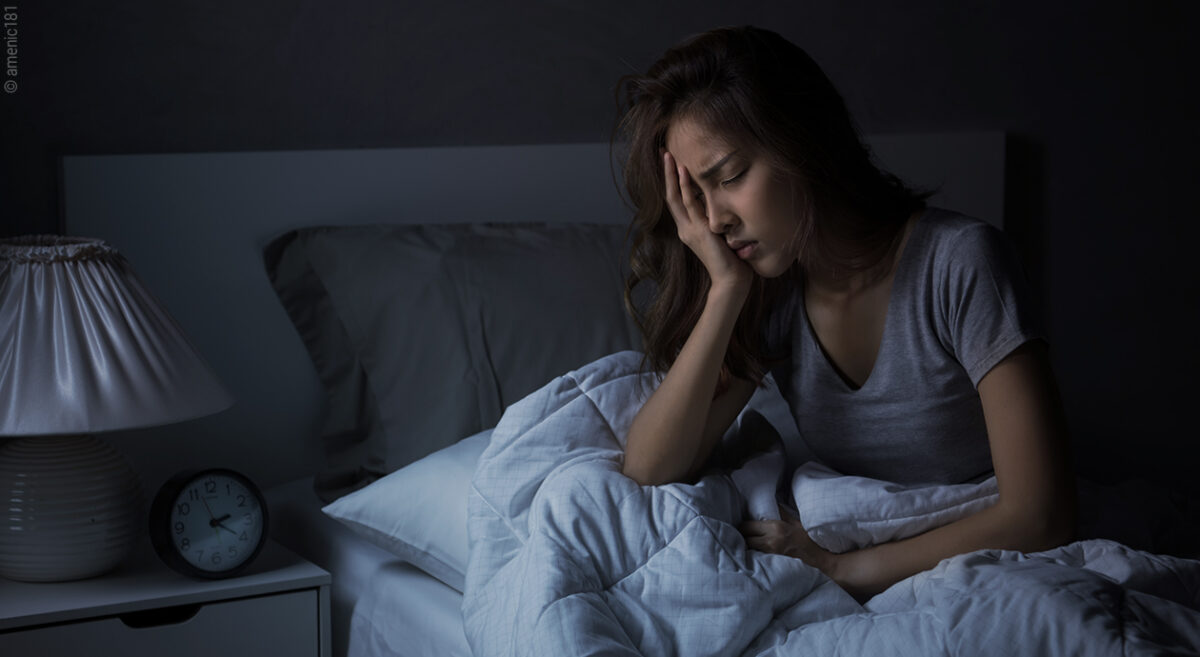 Titelbild zu Durchschlafstörungen: Eine Person sitzt im Bett, im dunklen Schlafzimmer und hält eine Hand vor das Gesicht. Der Gesichtsausdruck ist leidend.