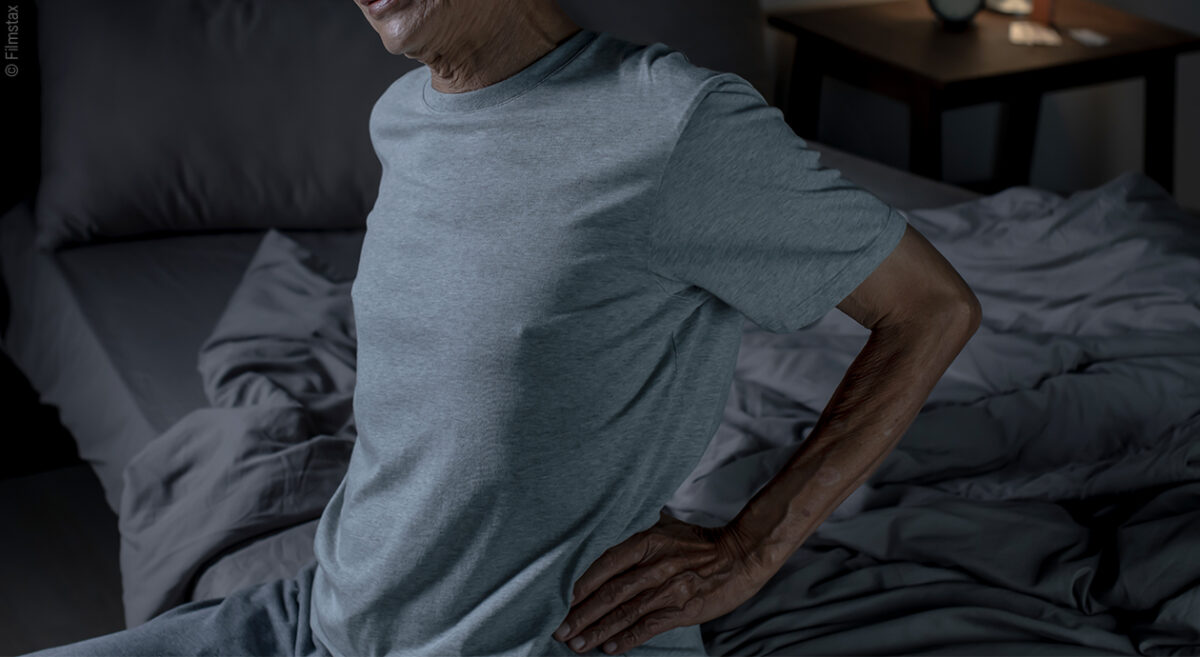 Foto: Eine Person sitzt am Rand eines Bettes und fasst sich an den unteren Rücken.