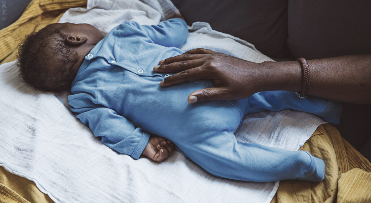 Ein Baby schläft auf dem Bauch, während die Hand einer erwachsenen Person auf seinem Rücken liegt.