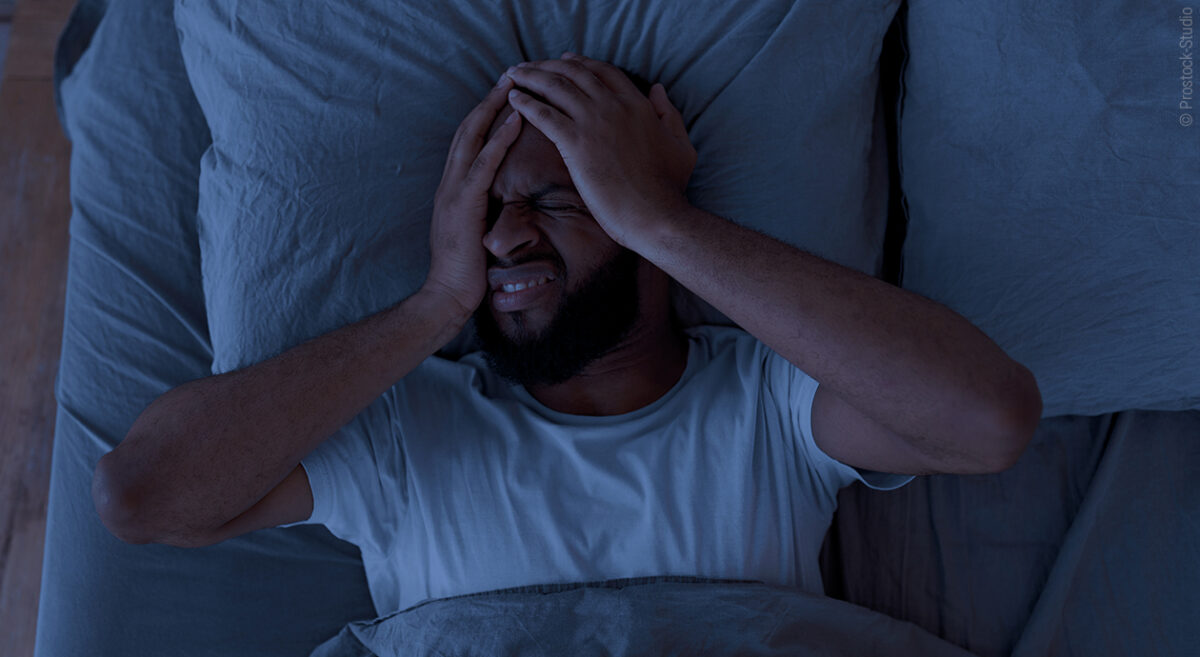 Foto: Eine Person liegt nachts im Bett und hält sich die Hände an den Kopf, das Gesicht sieht gequält aus.