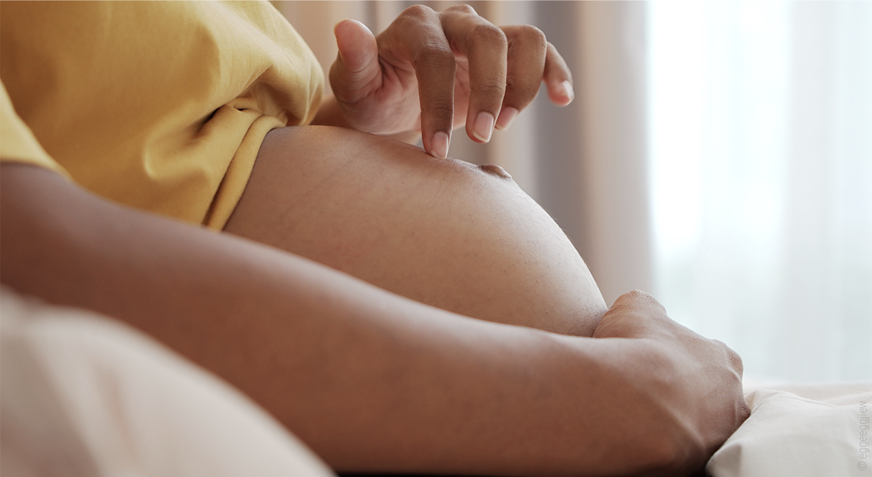 Foto: eine Person umfasst ihren schwangeren Bauch mit einer Hand von unten, die andere Hand tippt auf die Wölbung.