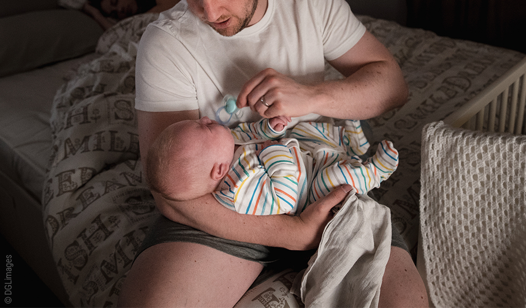 Foto: Eine Person sitzt auf einer Bettkante und hält ein Baby im einen, einen Schnuller im anderen Arm.