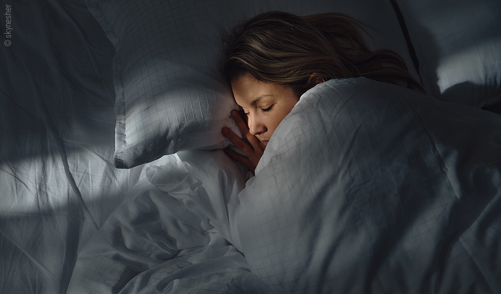 Foto: Eine Person liegt im Halbdunklen mit geschlossenen Augen im Bett. Etwas Licht fällt auf ihr Gesicht.