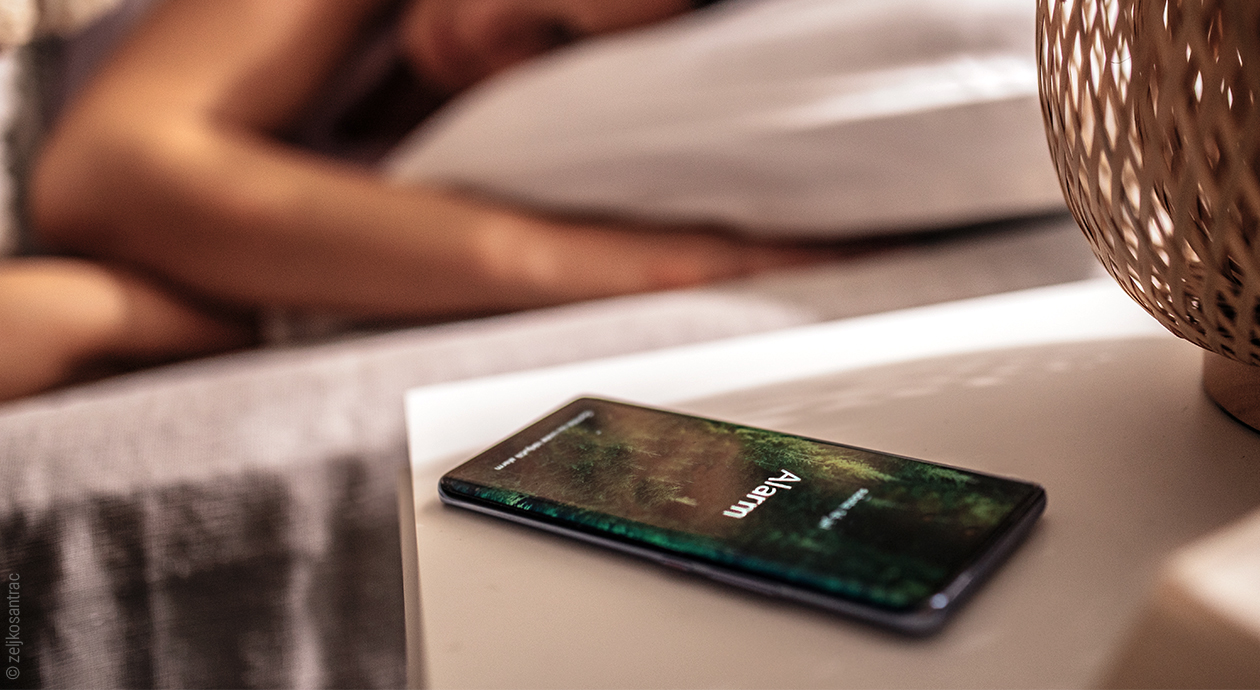 Titelfoto zum Thema Snooze-Wecker: Ein Smartphone auf der Tischkante zeigt Alarm, im Hintergrund liegt eine Person