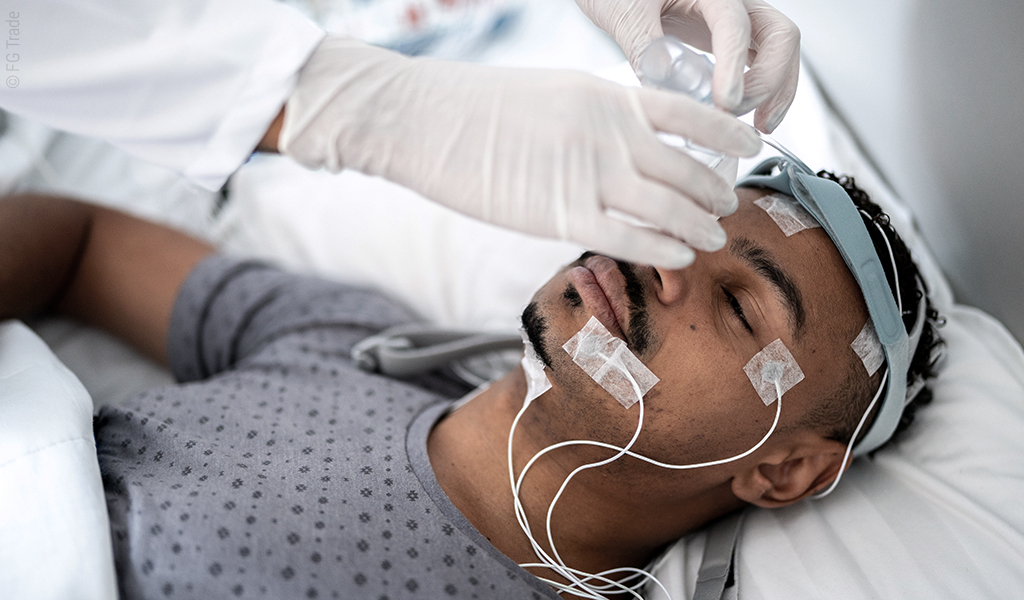Foto: Eine Person liegt im Bett, an ihrem Kopf sind Kabel und Elektroden befestigt