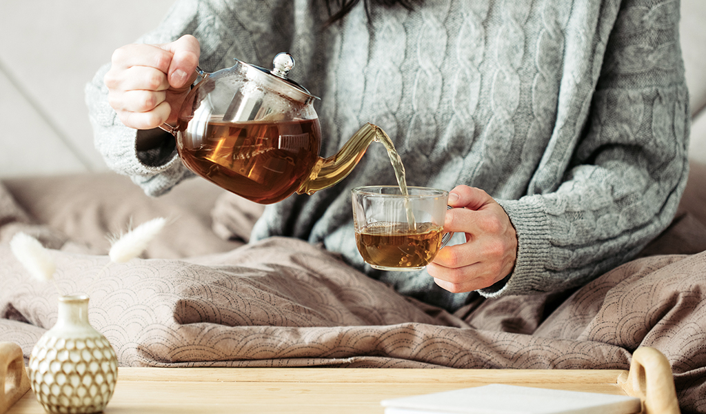 Foto: Eine Person ist in eine Decke gewickelt und gießt sich Tee aus einer Kanne ein