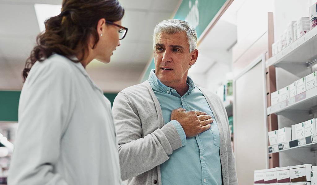 Foto: Eine Person in weißem Kittel spricht mit einem Kunden, der seine Hand an seine Brust hält, vor ihnen Medikamente in einem Regal
