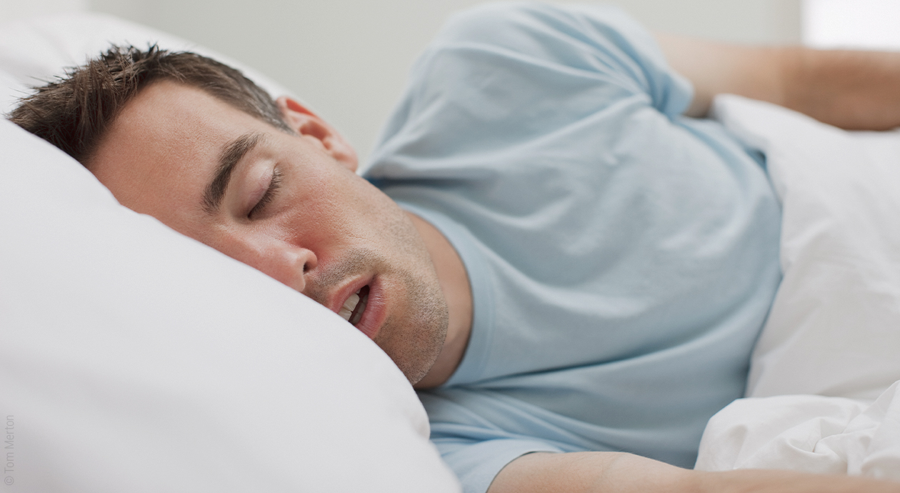Titelbild Sabbern im Schlaf: Eine Person liegt mit geschlossenen Augen und geöffnetem Mund auf der Seite im Bett.