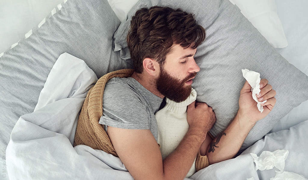 Foto: Eine Person liegt eingewickelt in eine Decke im Bett, hält eine Wärmflasche im Arm und ein Taschentuch in der Hand. 