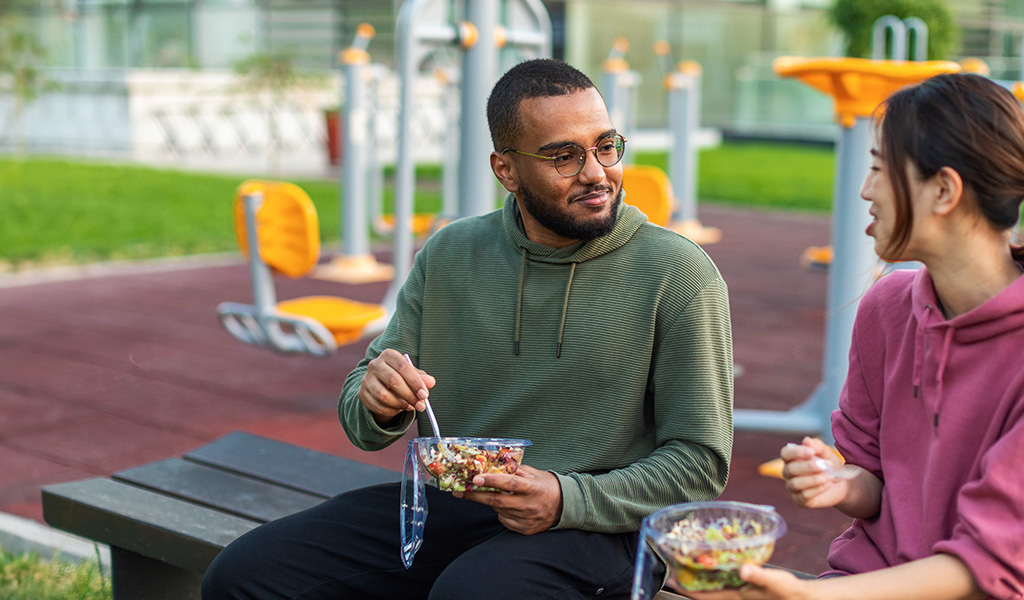 Foto: Zwei Personen mit Salatschüsseln auf dem Schoß sitzen einander zugewandt auf einer Bank im Freien.