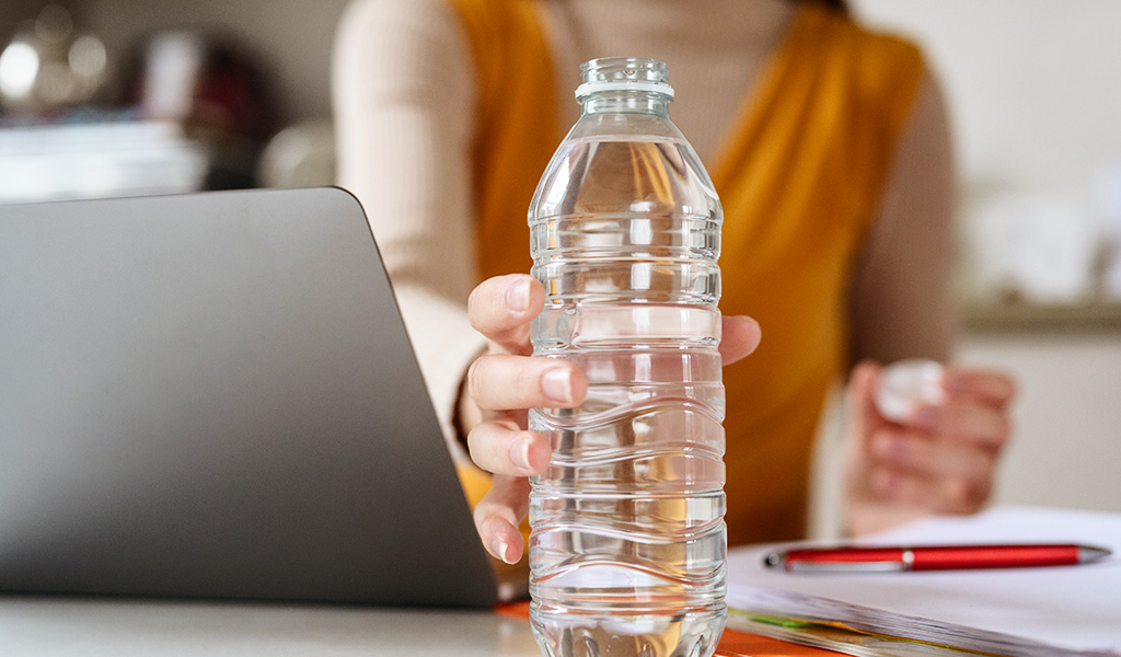 Foto: Eine Hand greift nach einer Flasche Wasser auf einem Schreibtisch.