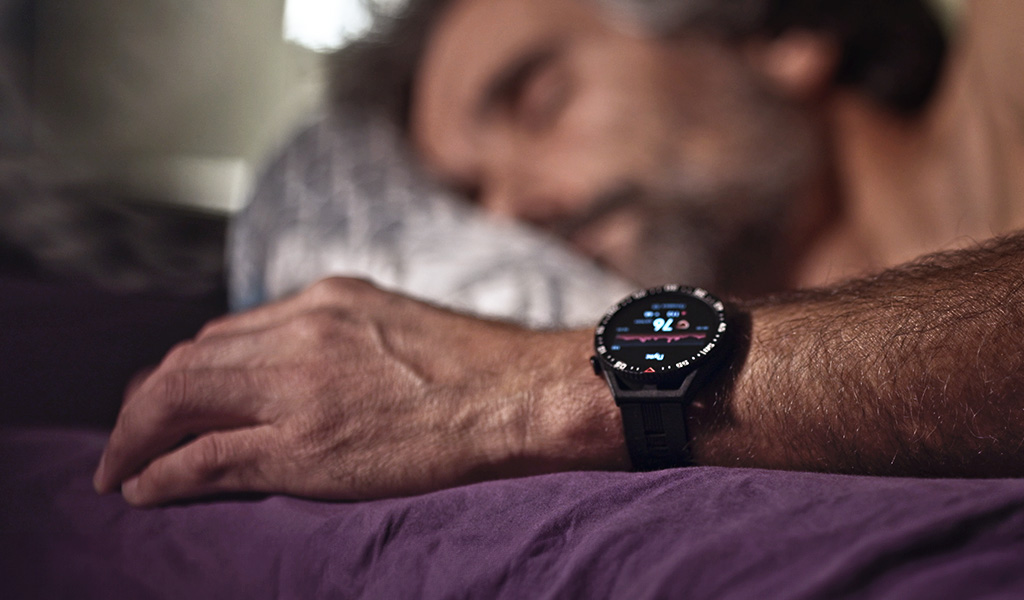 Foto: Fokus auf das Handgelenk einer im Bett liegenden Person, die eine Uhr trägt, auf der die Zahl 76 erleuchtet ist.