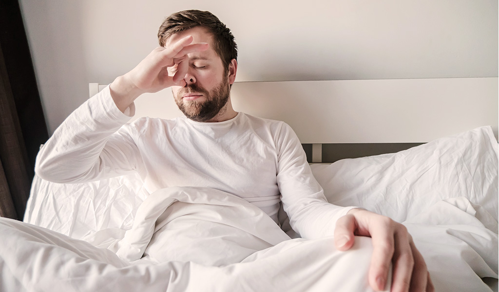 Foto: Eine Person sitzt halb zugedeckt im Bett und hält sich mit dem Daumen das rechte Nasenloch zu.